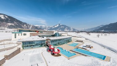 Aerial view of a winter hotel with indoor and outdoor swimming pools and water slide in a snowy landscape, surrounded by mountains. | © Alexander Maria Lohmann