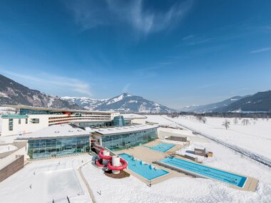 Aerial view of a winter hotel with indoor and outdoor swimming pools and water slide in a snowy landscape, surrounded by mountains. | © Alexander Maria Lohmann