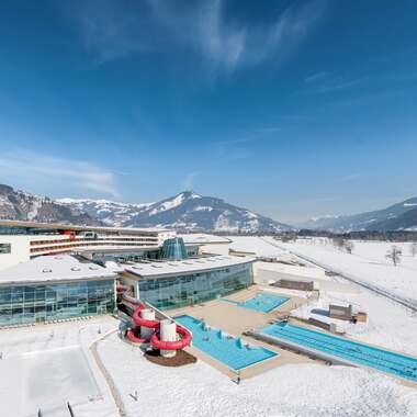 Aerial view of a winter hotel with indoor and outdoor swimming pools and water slide in a snowy landscape, surrounded by mountains. | © Alexander Maria Lohmann