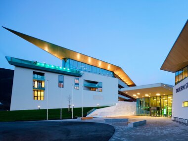 Modern building with a sloped roof and large entrance area, Tauern SPA in Kaprun at night. | © TAUERN SPA Zell am See-Kaprun