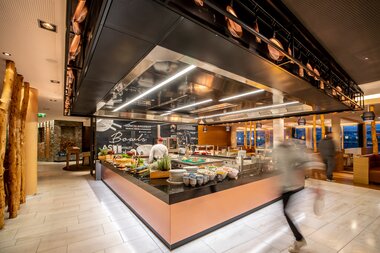 Interior of a hotel restaurant featuring a show kitchen and buffet, with light wood and black design elements. | © TAUERN SPA Zell am See-Kaprun