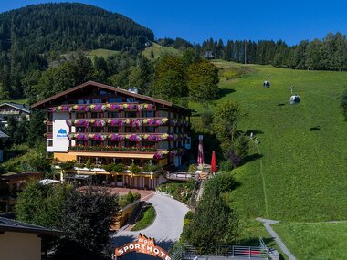 Sporthotel Alpin in a mountain landscape with green meadows and ski lifts in the background. | © Sporthotel Alpin Zell am See
