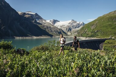 Two hikers exploring a lake landscape in the mountains with snow, a green hill, and a dam in the background. | © Zell am See-Kaprun Tourismus