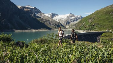 Two hikers exploring a lake landscape in the mountains with snow, a green hill, and a dam in the background. | © Zell am See-Kaprun Tourismus