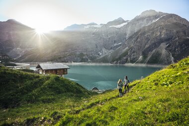 Two hikers by a high-altitude reservoir shore, surrounded by snow-capped mountains at sunrise. | © Zell am See-Kaprun Tourismus
