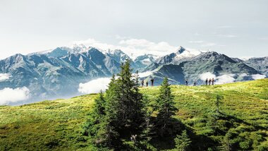 Panoramic view of the High Tauern with hikers on a green alpine, surrounded by mountains and clouds. | © Harry Liebmann 