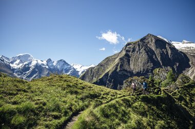 Trail through a green mountainous landscape with mountains in the background and hikers under a large rock | © Zell am See-Kaprun Tourismus