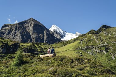 Two people sitting on a bench enjoying the view of green mountains and snowy peaks in the Kitzsteinhorn range. | © Zell am See-Kaprun Tourismus