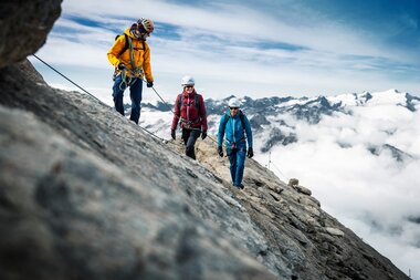 A group of climbers ascending a steep rock face amidst mountains, clouds, and peaks in the background. | © Zell am See-Kaprun Tourismus