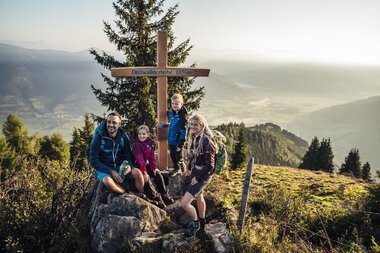 Family hiking on Maiskogel with a view of mountains and valley, next to a prominent summit cross. | © Zell am See-Kaprun Tourismus