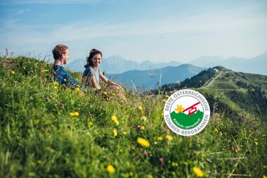 Two people sitting on a flowered meadow with a view of the Alps, on a sunny day at Schmittenhöhe.