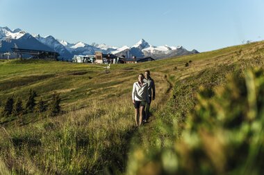 Two people hike on a green hillside with a view of mountains and a ski area on the Schmittenhöhe in Zell am See-Kaprun. | © Zell am See-Kaprun Tourismus
