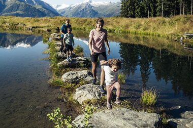 Family hiking on a stone walkway over a lake with mountains in the background on a sunny day. | © Zell am See-Kaprun Tourismus