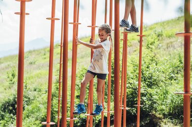 Children playing in Schmidolins Adventureworld on a climbing course with vertical poles, surrounded by nature. | © Schmittenhöhe