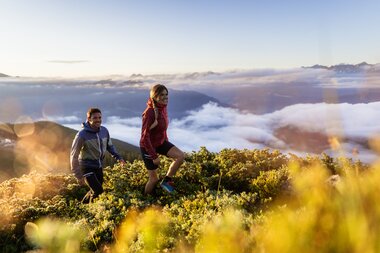 Two hikers on a mountain meadow at sunrise with clouds and mountains in the background. | © Branislav Rohal