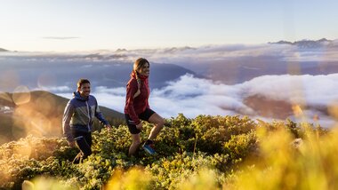 Two hikers on a mountain meadow at sunrise with clouds and mountains in the background. | © Branislav Rohal